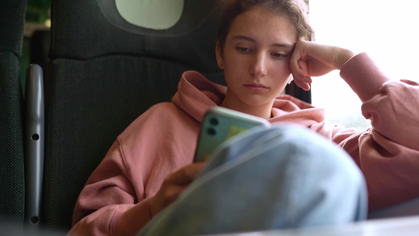 Attractive young teenager, in casual clothes, fashionable sweatshirt, jeans, sits near window in train car and enjoys trip, looks out window, chats on social networks on mobile phone. Slow motion Royalty-Free Stock Footage #1090963217