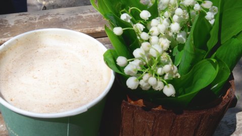4k video,static close up.lily of the valley flowers in cute little pot on wooden table outside,in a public park.cappuccino drink and white flowers, people in the background. sunny spring,summer day.