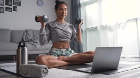 Sporty young Asian woman exercising at home, watching fitness video on Internet or having online fitness class, using laptop, living room interior.