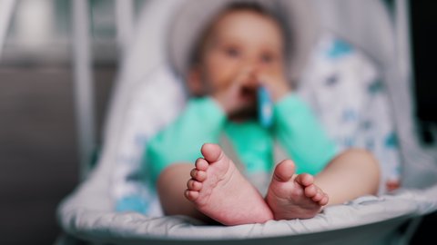 Little feet of a toddler in a baby chair. Active kid waving hands and legs in a swaying cradle. Blurred backdrop.