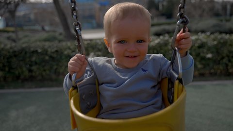 Cheerful boy rides swings on blurred background. Blond toddler enjoys activity at playground for children on sunny day closeup slow motion