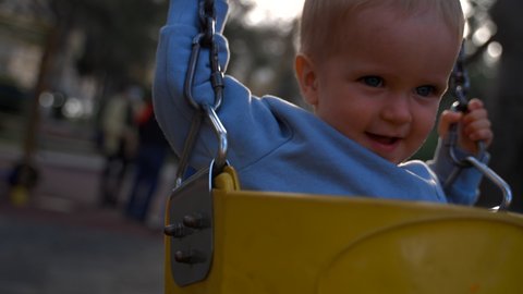 Toddler rides swings holding on to iron chains on blurred background. Blond boy enjoys sunny day at playground in city park closeup slow motion