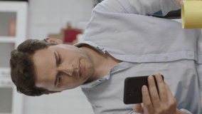 Smiling young man using smartphone and drinking coffee at home.Young user sipping coffee while browsing social media accounts on smartphone, listening to speaker talking behind camera.