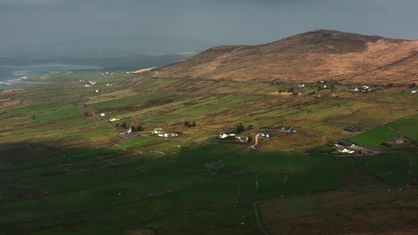 Loher Ringfort, Kerry, Ireland, March 2022. Drone orbits the ancient monument from the southwest as clouds cast shadows across the landscape with Ballinskelligs Bay and Atlantic Ocean in the distance.
