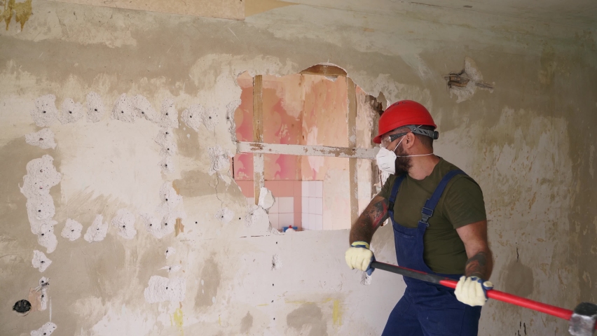 Contractor wrecks wall with sledgehammer making hole for rearrangement. Man doing manual dismantling and demolition works with big hammer hits for apartment renovation. Construction worker in uniform.