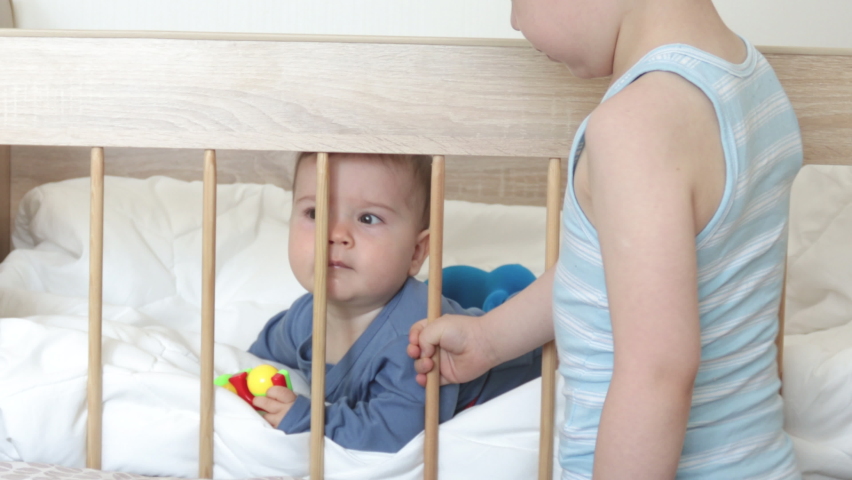 4k,kid is swinging a crib with baby boy inside.big brother swinging infant in wooden bed to fall asleep.toddler puts tongue on wooden bars,refuse to sleep.2 brothers,child's room. Royalty-Free Stock Footage #1091005229