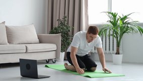 redhead man in white t-shirt works out in the living room. Man doing sports on a mat watching videos on a laptop in the living room with water and dumbbells on the floor