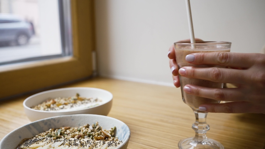 Female hands take glass of fresh fruit smoothie with straw from wooden table. Sweet vegan bowls placed on table near window closeup
