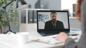 Close up of woman sitting at office desk and taking notes during video meeting with her bearded mature boss. Focus on laptop screen with caucasian man in suit.