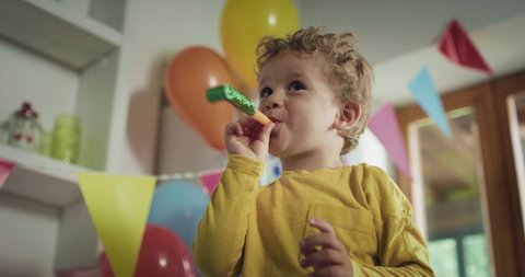 Cinematic authentic shot of cute happy little toddler boy is blowing party trumpet and smiling in camera during birthday celebration with family at home. 