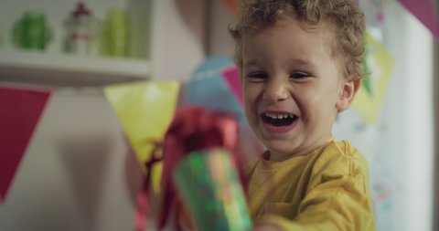 Cinematic authentic shot of cute happy little toddler boy is blowing party trumpet and smiling in camera during birthday celebration with family at home. 