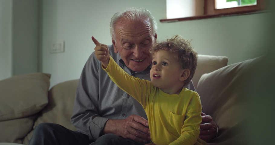 Cinematic shot of happy senior grandfather and cute toddler baby boy having fun to watch television together for family entertainment on sofa in living room at home.