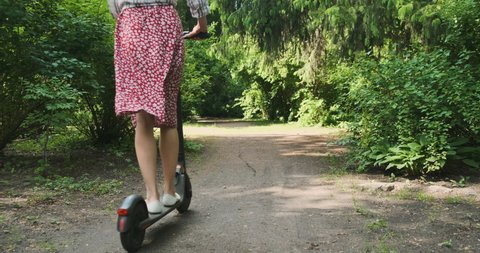 young woman rides an electric scooter along a path in a park among green trees. Rear view, daytime, wide shot.