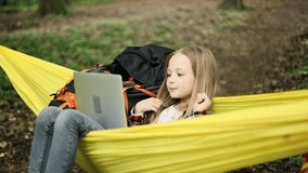 Cute little girl sitting in a hammock in the park and talking on laptop by video chat. Child relax in a yellow hammock.