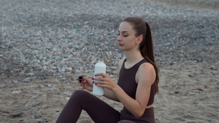 A beautiful girl drinks water from a bottle on the beach.