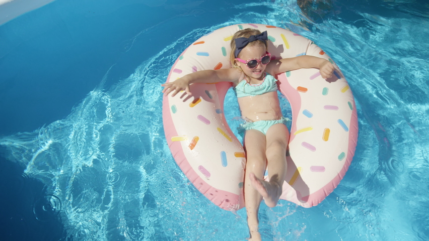 TOP DOWN: Adorable girl with sunglasses on a floatie donut in blue swimming pool. Cute girl having fun floating and making water splashes with her legs. Cheerful kid enjoying summer in backyard pool. Royalty-Free Stock Footage #1091017557