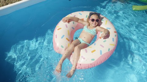 TOP DOWN: Adorable girl with sunglasses on a floatie donut in blue swimming pool. Cute girl having fun floating and making water splashes with her legs. Cheerful kid enjoying summer in backyard pool.