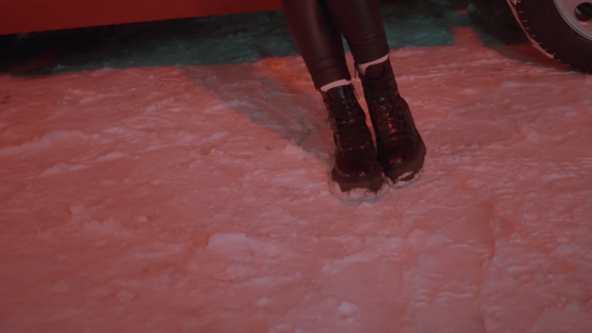 Woman in tight black leather leggings, coat and high boots stands still on snow, legs crossed, hands and body lean on red vintage convertible car, snowfall in winter night, slow zoom, tilt up close-up