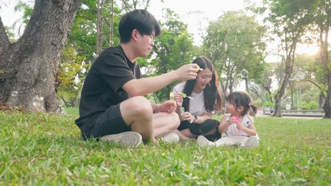 BKK, Thailand- Apr 22, 2022 Asian young adult parents playing with cute baby girl toddler by blowing soap bubbles sit down relaxing in the park under trees and sunset, Happy Asian family enjoy outdoor