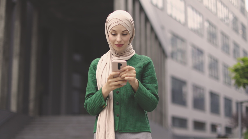 Low angle view of beautiful muslim woman standing near modern office building with smartphone in hands. Young business lady in formal wear and hijab checking work emails outdoors. | Shutterstock HD Video #1091028893