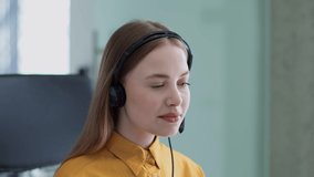 Young positive professional woman helpline operator wearing headset turning face to camera and smiling, sitting at workplace, slow motion