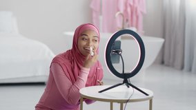 Product review. Young positive black muslim lady wearing headscarf applying hygienic lipstick on her lips, recording video for her beauty blog on smartphone, slow motion