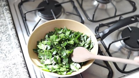 Female hands cooking ora-pro-nobis in frying pan. Pouring oil and wooden spoon. Pereskia aculeata is a popular vegetable in parts of Brazil
