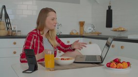 Lovely positive adult female in casual clothes enjoying muesli breakfast with fresh sliced fruits and berries, watching video content online using laptop while spending leisure in domestic kitchen.
