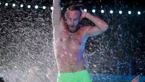 Happy homosexual man celebrating gay pride with friends at night LGBTQ pool party in a private villa swimming pool. Bisexual male in swimwear dancing splashing water in luxury resort. Slow motion.