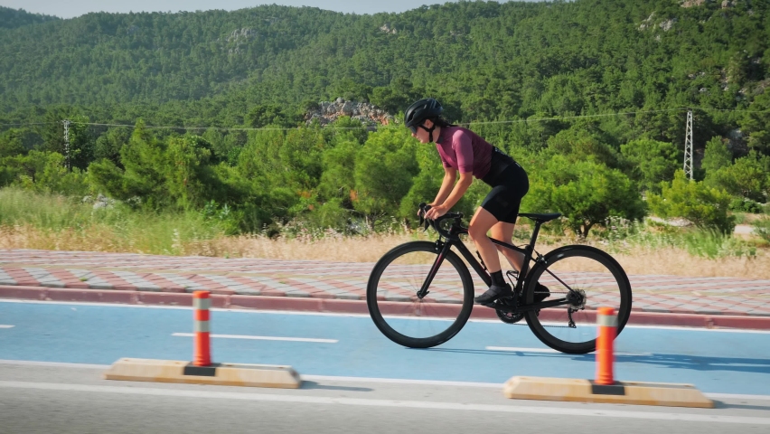 Cycling training. Young brunette female cyclist in black helmet and cycling apparel riding bicycle on bicycle lane in sunny morning, preparing for bike race season, doing hard intense trainings