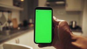 Man holding mobile phone with green screen at night in kitchen. Mock up chroma key surfing internet, watching content video.
