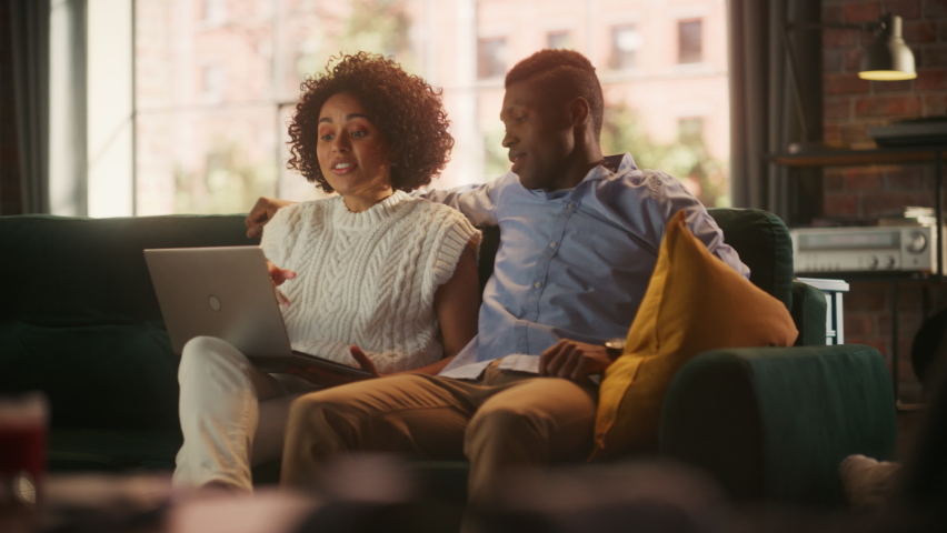 Cute Black Couple in Love Use Laptop Computer, Shopping on Internet, Using Social Media. Boyfriend and Girlfriend Have Fun, Laughs, while Sitting on the Couch in the Cozy Apartment. Female Empowerment