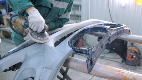 Worker in gloves and jumpsuit cleans surface of auto bumper with sanding machine professionally in car service garage before painting closeup