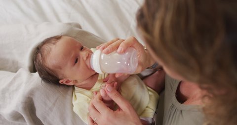 Video of happy caucasian mother feeding newborn baby with bottle on bed. motherhood, parental love and taking care of newborn baby concept digitally generated video.