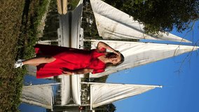 Luxury travel vacation. Vertical video of elegant woman in red dress walking near docked sailing yacht against the backdrop of the sea, sunset and nature.