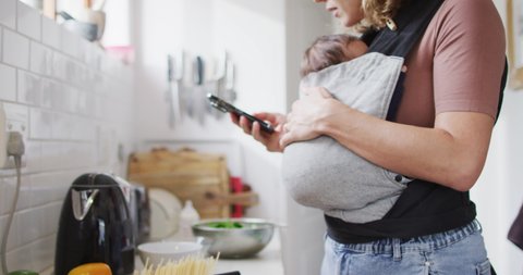 Video of caucasian mother with newborn baby in baby carrier using smartphone in kitchen. motherhood, parental love and taking care of newborn baby concept digitally generated video.