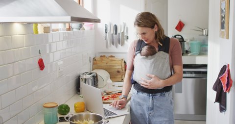 Video of caucasian mother with newborn baby in baby carrier using laptop in kitchen. motherhood, parental love and taking care of newborn baby concept digitally generated video.