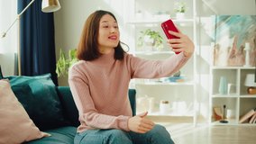 Asian smiling woman talking on video call. Young Korean female student speaking on smartphone, using virtual conference, sitting on sofa in living room. Communicating online via internet.