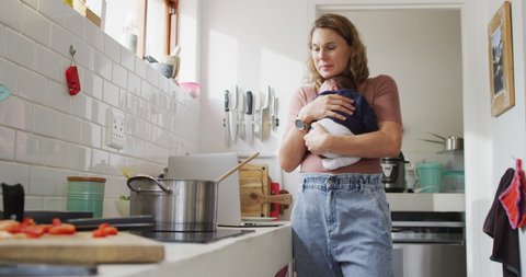 Video of caucasian mother with newborn baby in baby carrier cooking in kitchen. motherhood, parental love and taking care of newborn baby concept digitally generated video.