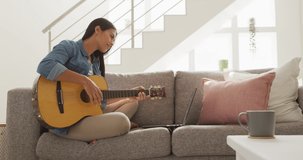 Video of happy biracial woman sitting on sofa and playing guitar. Lifestyle, music, hobby and spending free time at home concept.