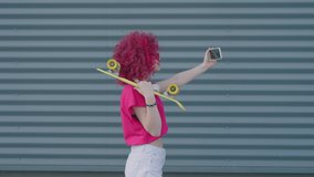 A pink curly teenager is recording video blog with smartphone outdoors. Hipster girl is creating content for social media account. Blogging or internet and video call or communication concept.