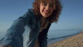 Smiling woman video chatting with friend on smartphone. Joyful girl waving hands at camera on beach. Excited lady showing sea landscape by video call. Woman feeling happy at vacation. Online concept