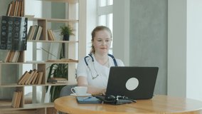 Female doctor in white coat with stethoscope around her neck is working at clinic with laptop. general practitioner drinks coffee while working. Telemedicine, concept of remote medical care.