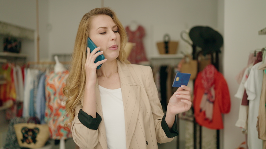 Young blonde woman customer talking on the smartphone holding credit card at clothing store