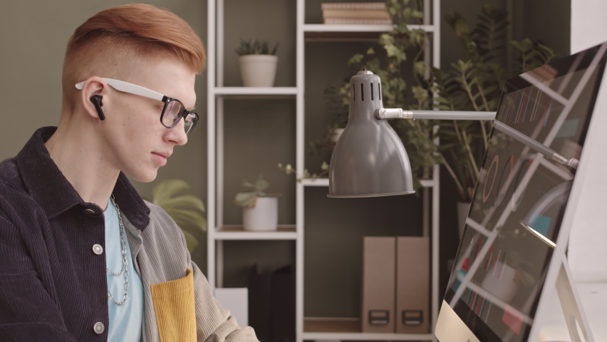 Waist up slowmo of young Caucasian ginger haired man with mono headset in his ear working on computer in office while his female colleague analyzing financial data in background