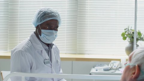 Rack focus shot of African American doctor in protective mask, hat, gloves and lab coat sitting at bedside in hospital ward and speaking with elderly Caucasian patient with nasal cannula