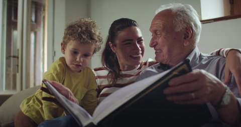 Cinematic close up shot of happy family: grandfather, daughter and grandson toddler boy are having fun looking at photo album with life memories together for family entertainment on sofa at home.