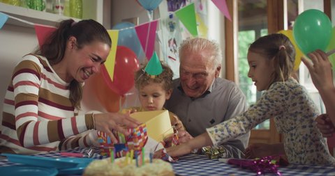 Cinematic authentic shot of cute happy little toddler boy wearing party hat is having fun to open his birthday present during celebration with family at home.