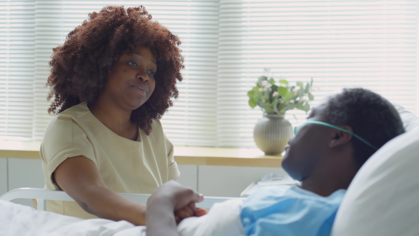 Positive African American woman sitting at bedside in hospital ward, holding hand of sick husband, chatting with him and laughing during visit