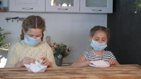 Two children in protection mask play at home.girls are sitting at home in quarantine, making paper boats. Childhood dream of playing paper boats. mask protects breath of children. Protective face mask